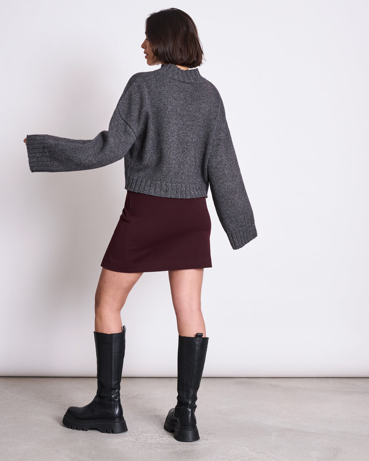 A woman with short brown hair faces away, wearing a loose gray EcoVero™ sweater, JAN-N-JUNEs MINI SKIRT AINOHA in intense rust, and tall black boots. She poses with one arm slightly raised in a minimal, modern setting.