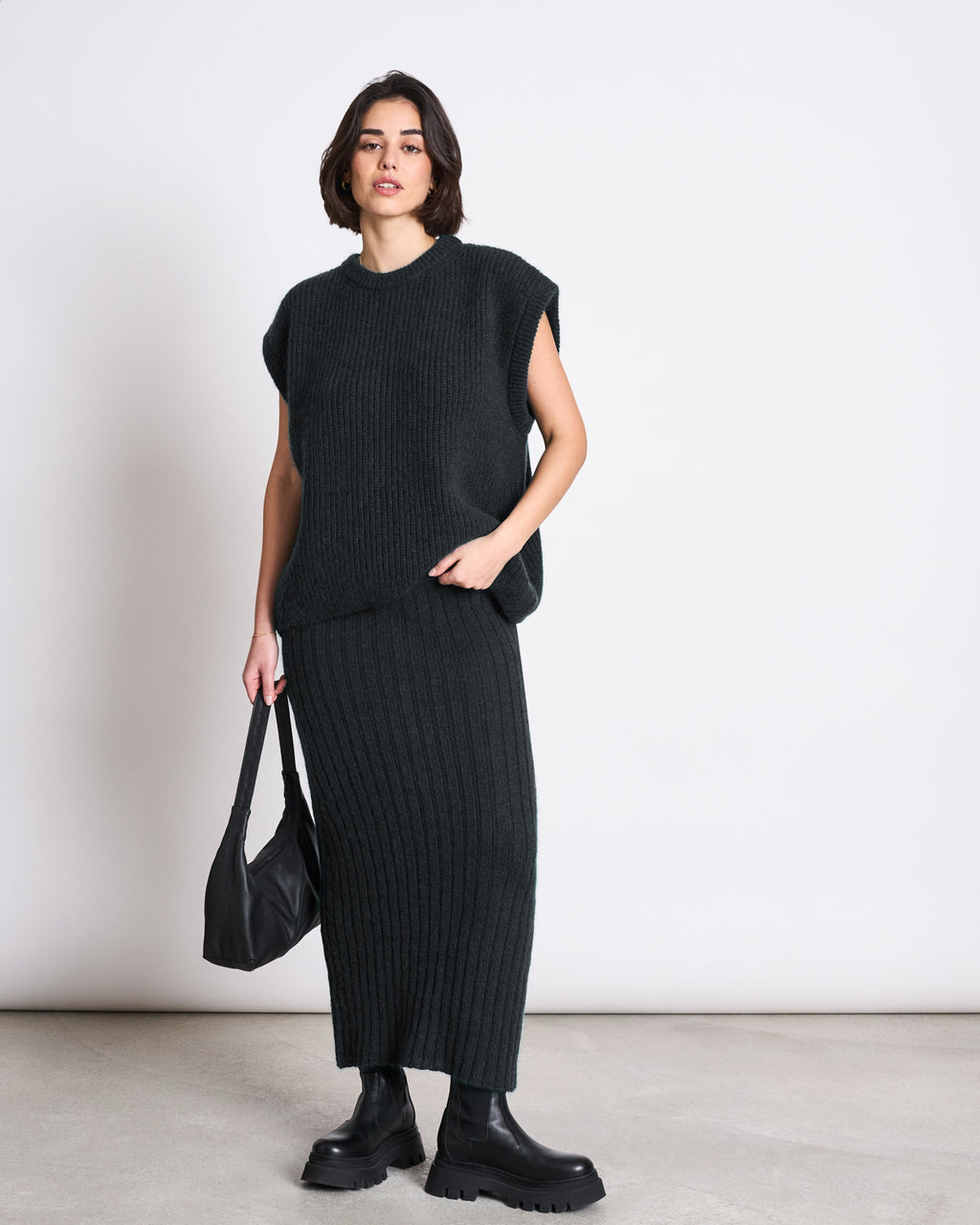 A woman stands against a plain light background wearing the jan-n-june WOOLY SLIPOVER THANDA BLACK, an oversized sleeveless ribbed knit top paired with a matching long skirt, styled with black chunky boots and a black handbag.