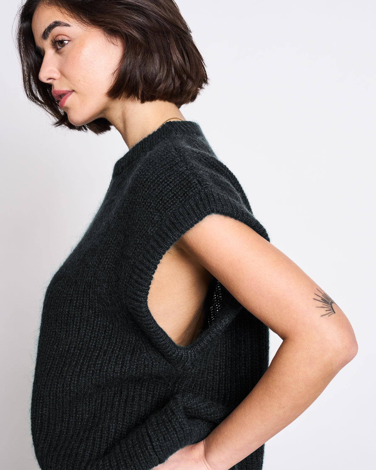 A woman with short dark hair stands in profile, wearing the jan-n-june WOOLY SLIPOVER THANDA BLACK in an oversized fit. Her left hand rests on her hip, highlighting a sunburst tattoo. The background is plain and light-colored.