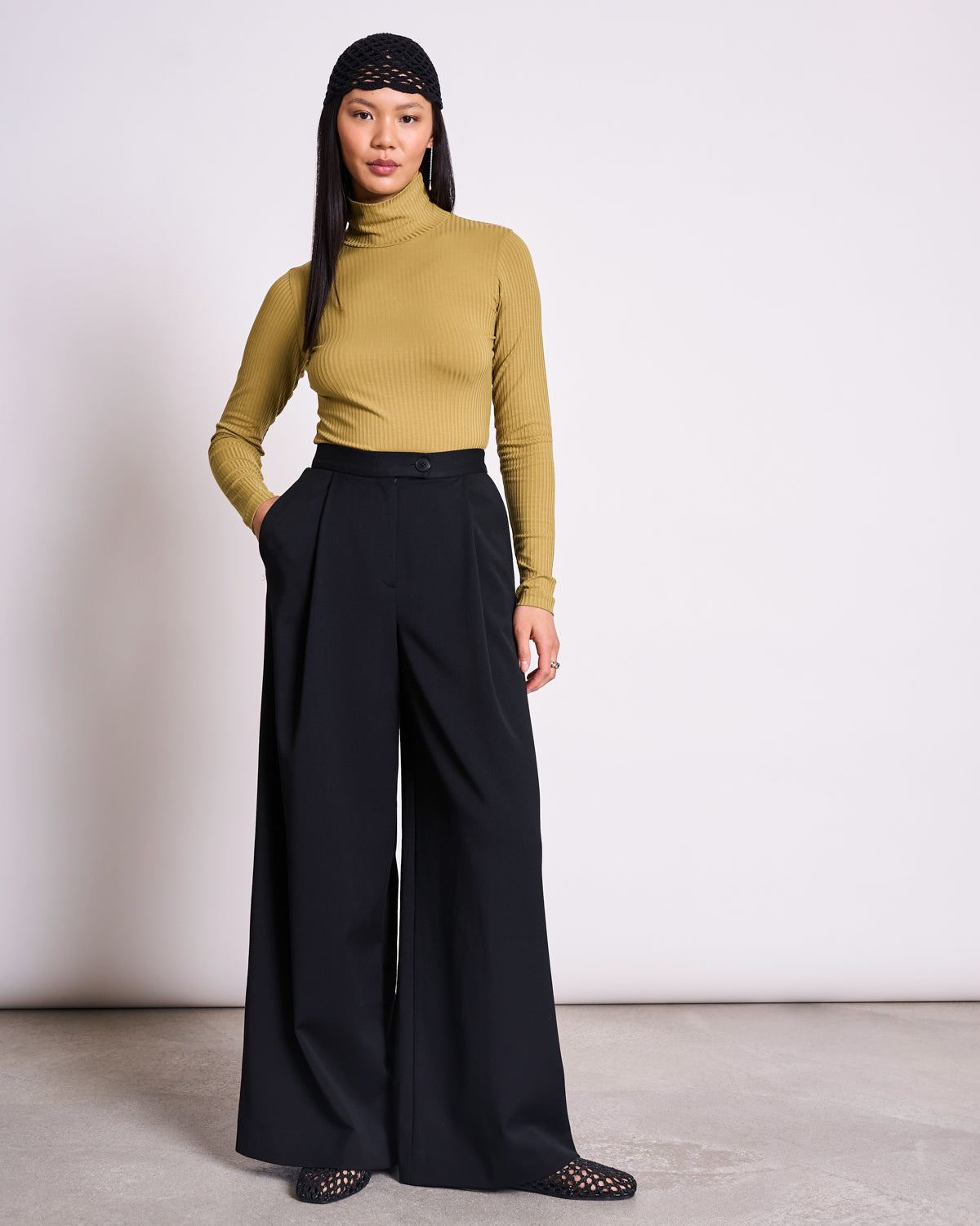 A woman stands against a plain light background, wearing the jan-n-june TENCEL™ LYOCELL RIB TURTLENECK in Mio Woody Olive. She pairs it with black wide-leg trousers, netted shoes, and a black headscarf, gazing confidently at the camera.