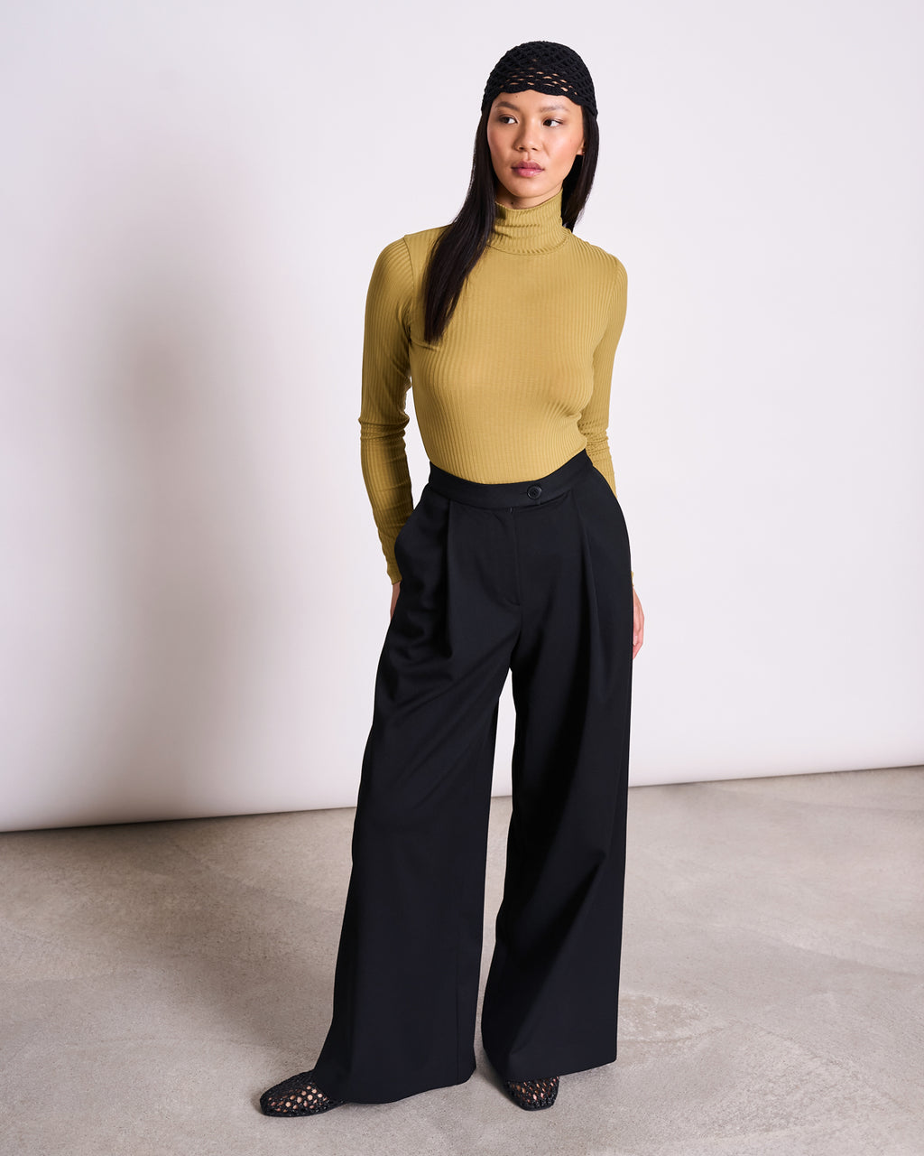 A woman wears the jan-n-june TENCEL™ LYOCELL RIB TURTLENECK in MIO WOODY OLIVE, paired with wide-leg black trousers, a black netted headscarf, and black mesh shoes. She stands on a light floor, looking slightly left with a neutral expression.