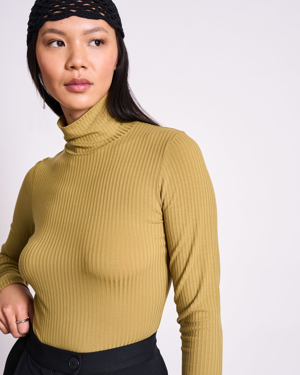 A woman in the jan-n-june TENCEL™ LYOCELL Rib Turtleneck MIO in Woody Olive and black pants stands against a light background, looking sideways. She has long dark hair and wears a black knit headband.
