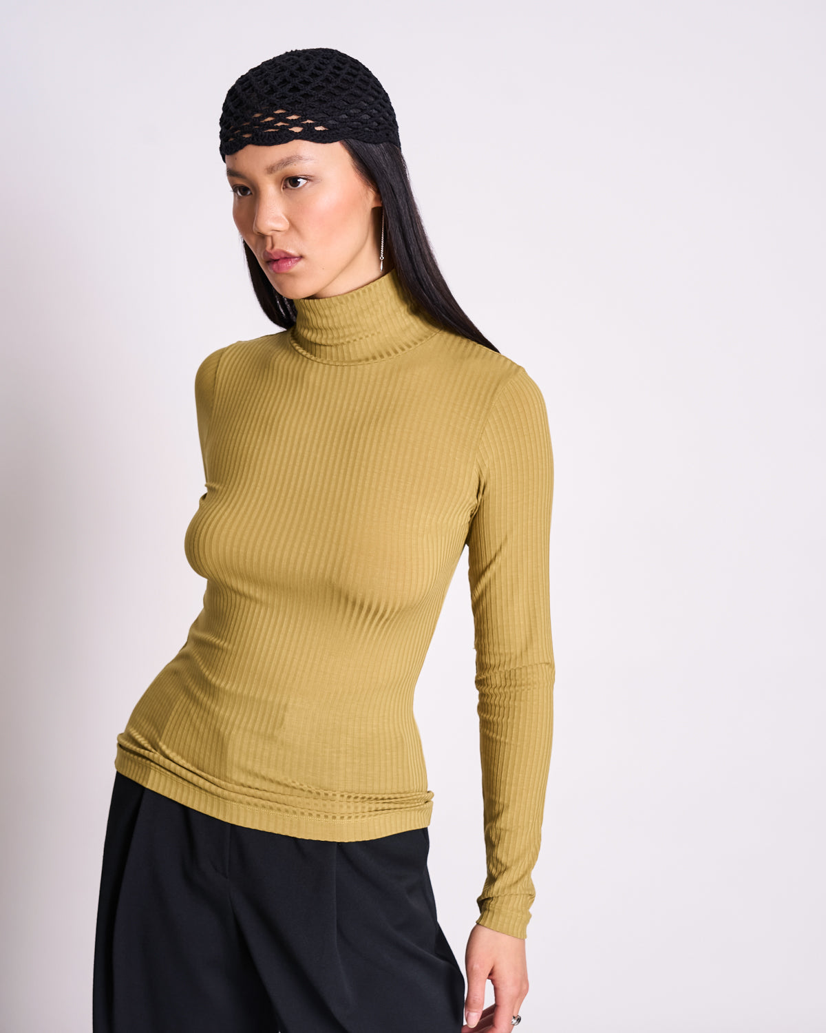 A woman stands against a plain background in jan-n-june’s form-fitting TENCEL™ LYOCELL rib turtleneck in woody olive and black pants, wearing a black mesh head covering.