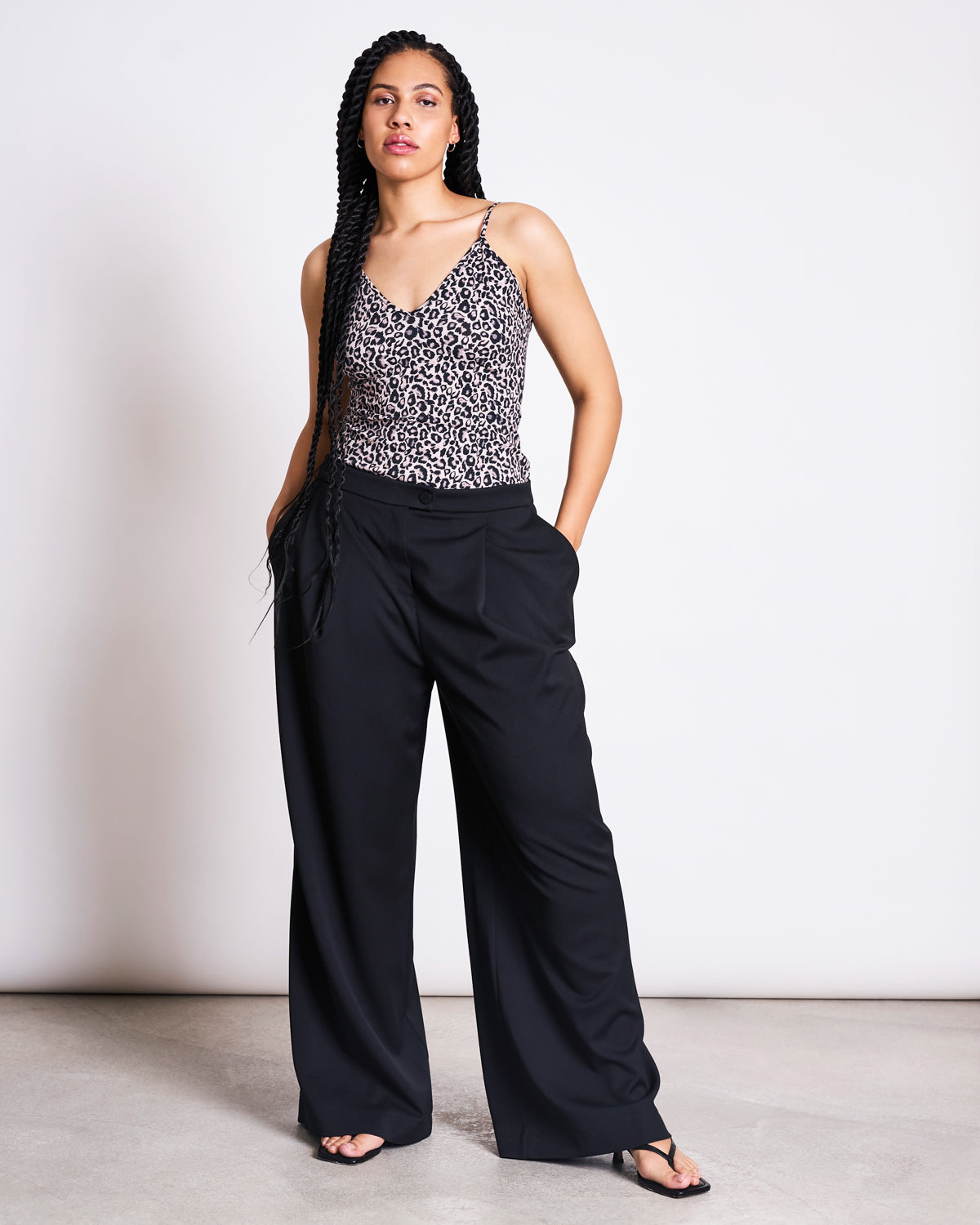 A woman with long braids wears the jan-n-june TOP MATILDA LEO GOTS, paired with black wide-leg pants and black sandals. She stands confidently with her hands in her pockets against a plain white background.
