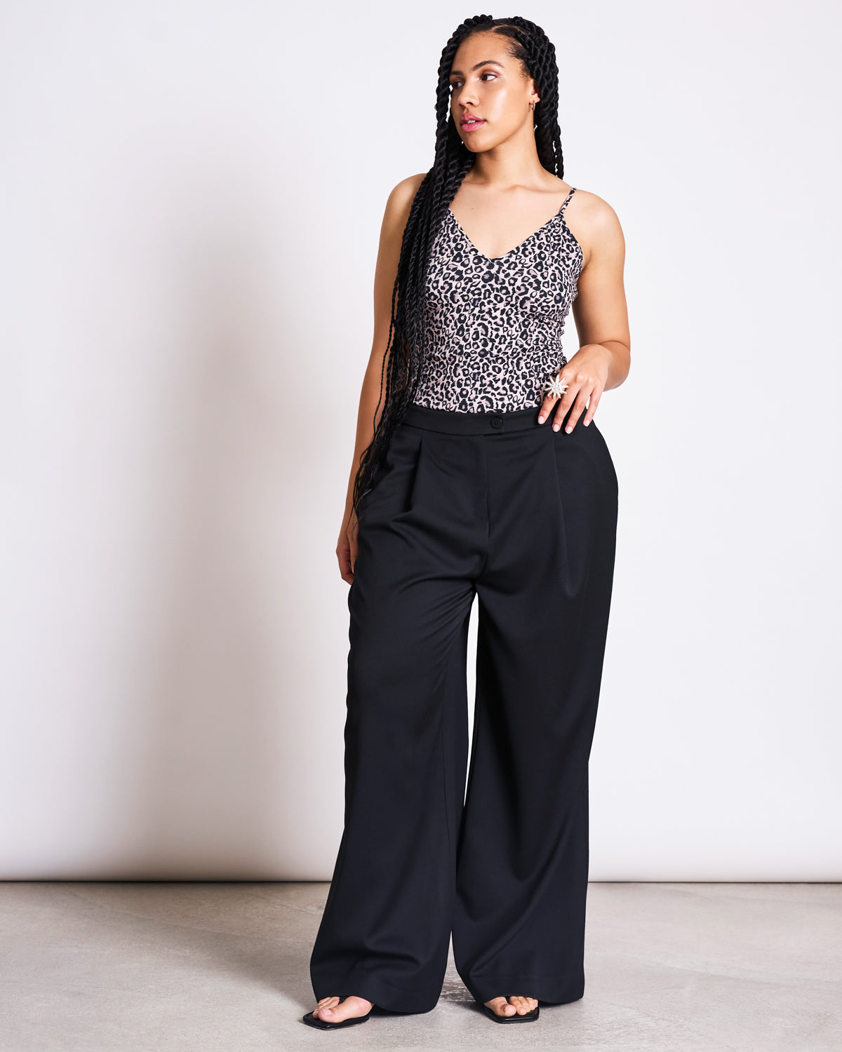 A woman with long braids wears the jan-n-june TOP MATILDA LEO GOTS, a sleeveless leo print top, paired with high-waisted black wide-leg trousers. She stands against a plain light background, looking to the side with one hand in her pocket.
