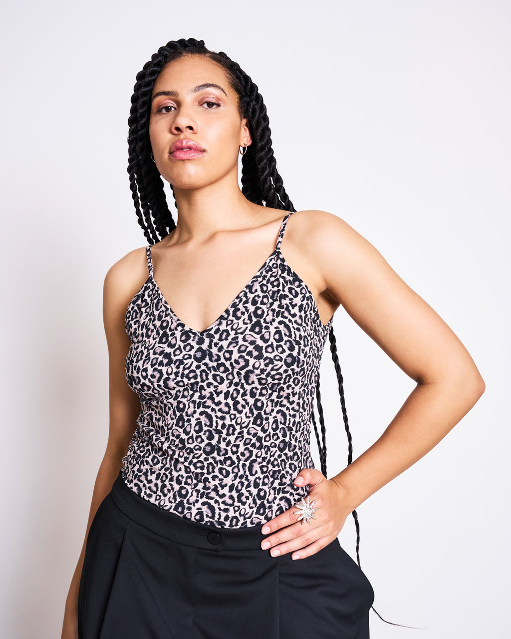 A woman with long braided hair models the jan-n-june TOP MATILDA LEO GOTS, a leo print spaghetti strap top, paired with black pants. She stands confidently against a plain white background, one hand on her hip and a neutral expression.
