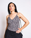 A woman with long braided hair models the jan-n-june TOP MATILDA LEO GOTS, a leo print spaghetti strap top, paired with black pants. She stands confidently against a plain white background, one hand on her hip and a neutral expression.