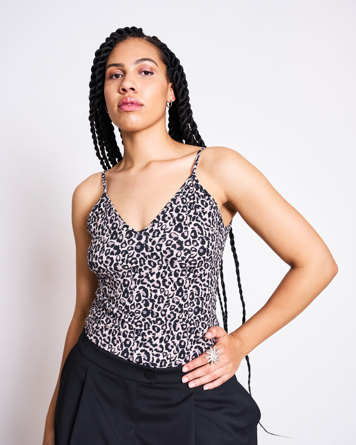 A woman with long braided hair models the jan-n-june TOP MATILDA LEO GOTS, a leo print spaghetti strap top, paired with black pants. She stands confidently against a plain white background, one hand on her hip and a neutral expression.