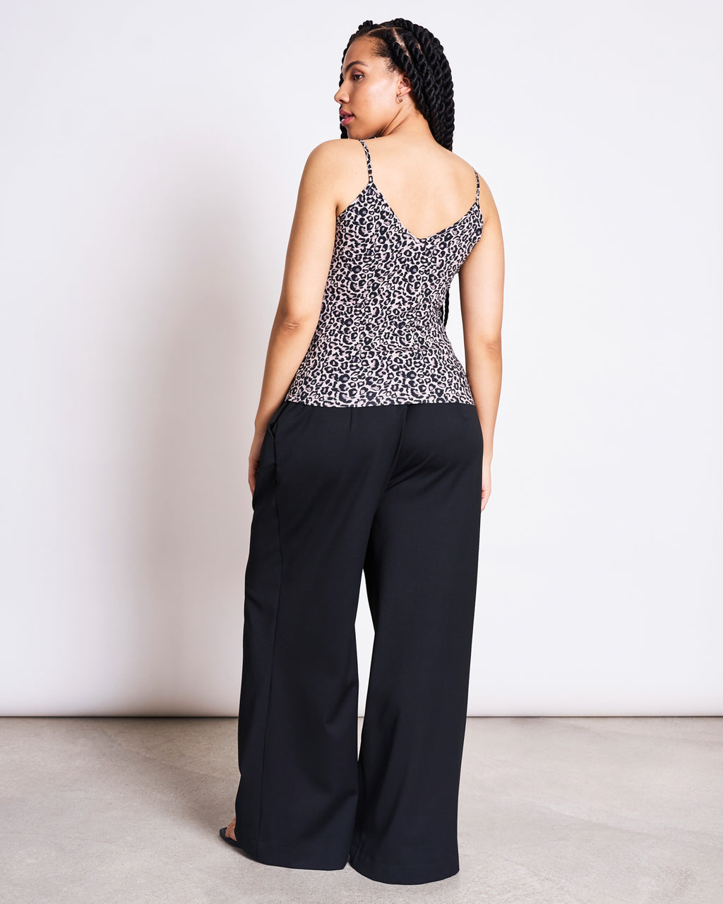 A woman with braided hair stands facing away, wearing the jan-n-june TOP MATILDA LEO GOTS and loose black pants, set against a plain indoor background.