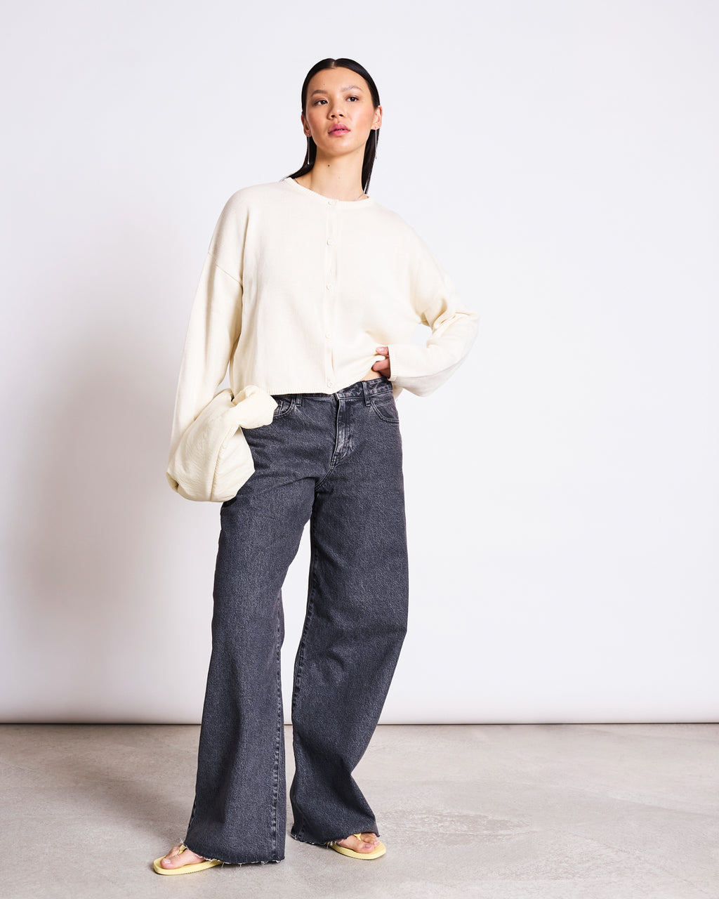 A woman with straight dark hair poses against a white background, wearing the jan-n-june LIGHT CARDIGAN ASAS OFFWHITE GOTS, paired with high-waisted wide-leg dark gray jeans and yellow open-toe sandals for a sustainable look.