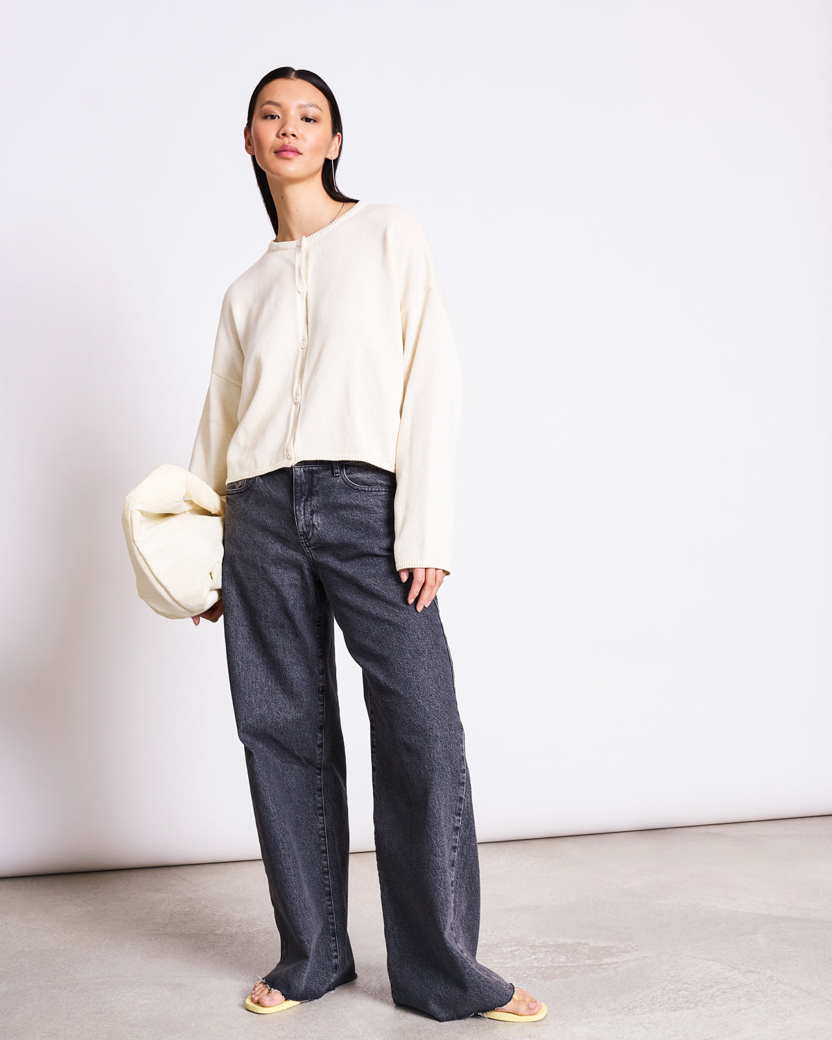 A woman stands against a white wall wearing the jan-n-june LIGHT CARDIGAN ASAS OFFWHITE GOTS, paired with loose dark gray jeans and yellow sandals. She confidently holds a matching cream clutch, showcasing sustainable style.