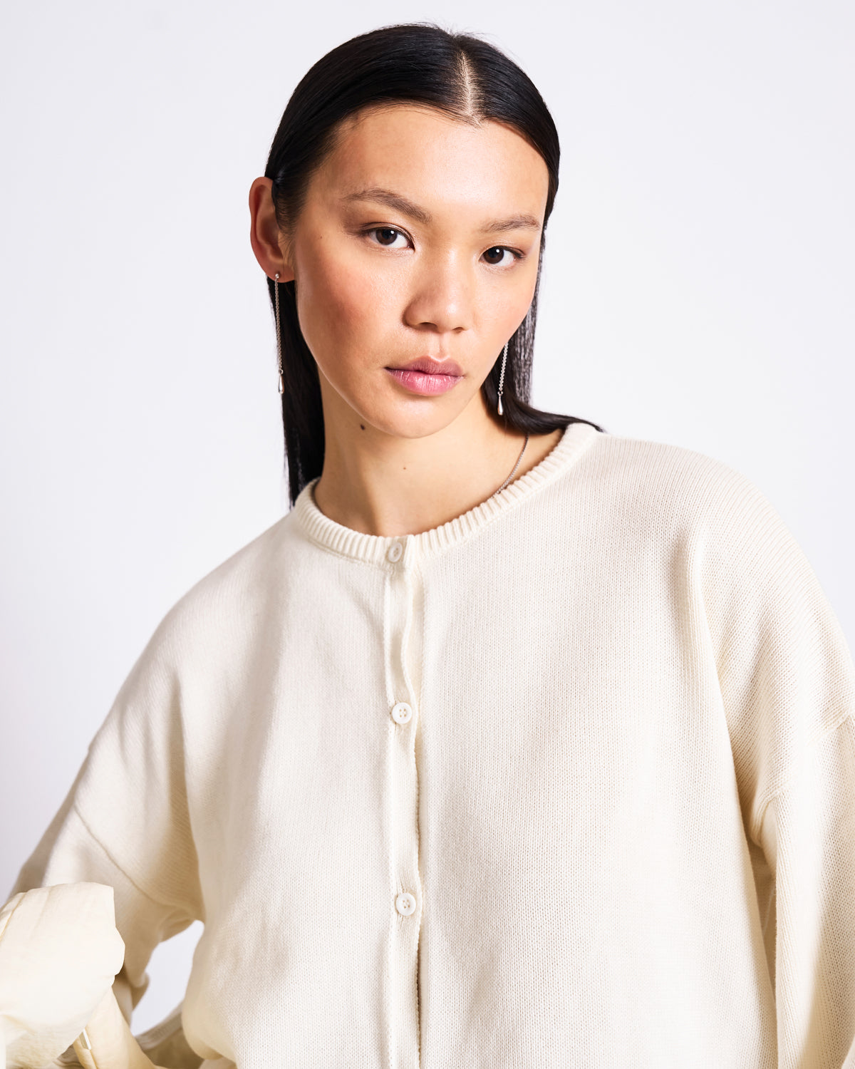 A woman with straight dark hair and natural makeup wears the jan-n-june LIGHT CARDIGAN ASAS OFFWHITE GOTS, an oversized cream organic cotton cardigan, standing against a plain white background and looking at the camera.