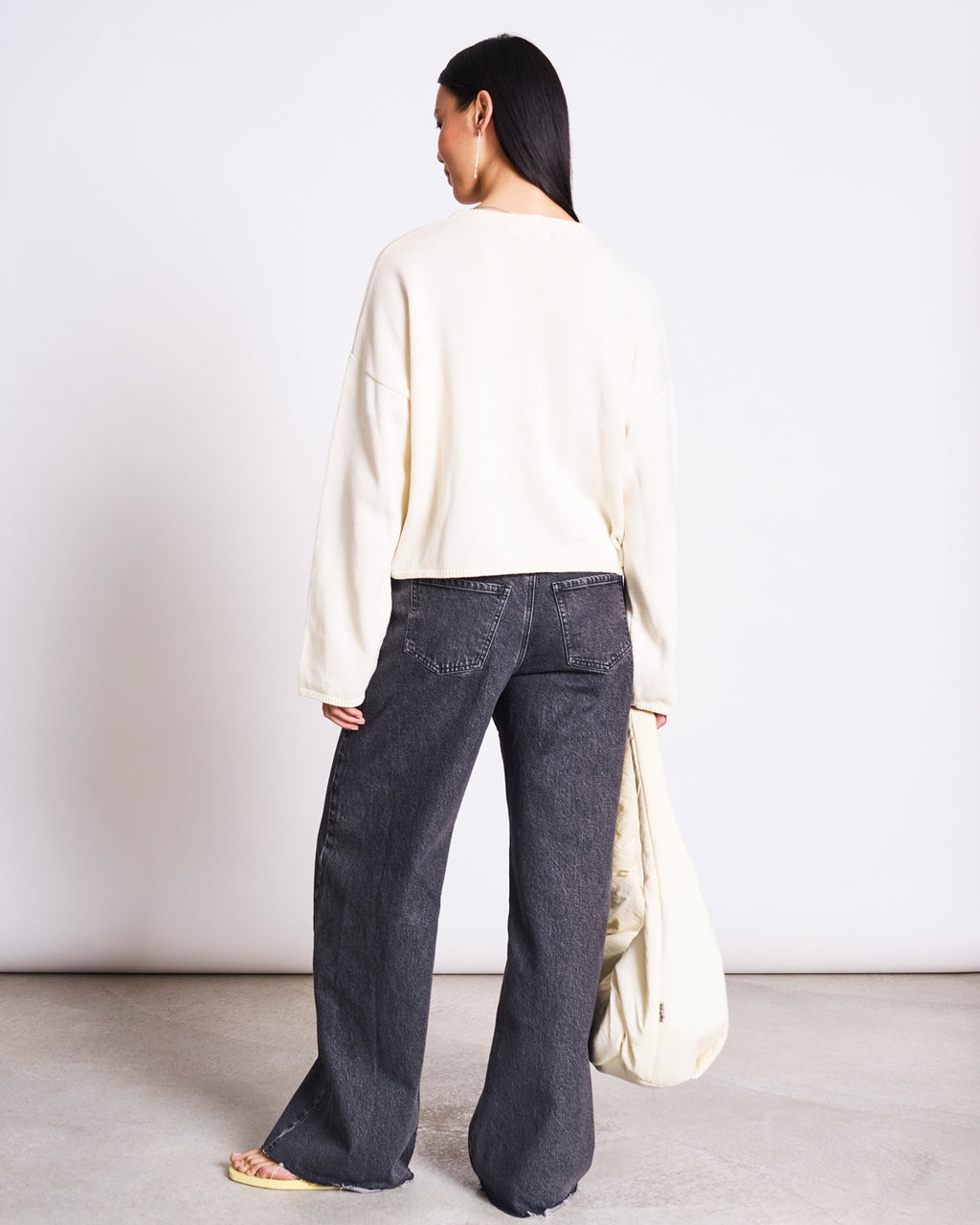 A woman faces away indoors, wearing the jan-n-june LIGHT CARDIGAN ASAS OFFWHITE GOTS with wide-leg dark gray jeans and yellow sandals. She carries a light tote bag against a plain, minimalistic background that highlights sustainable fashion.