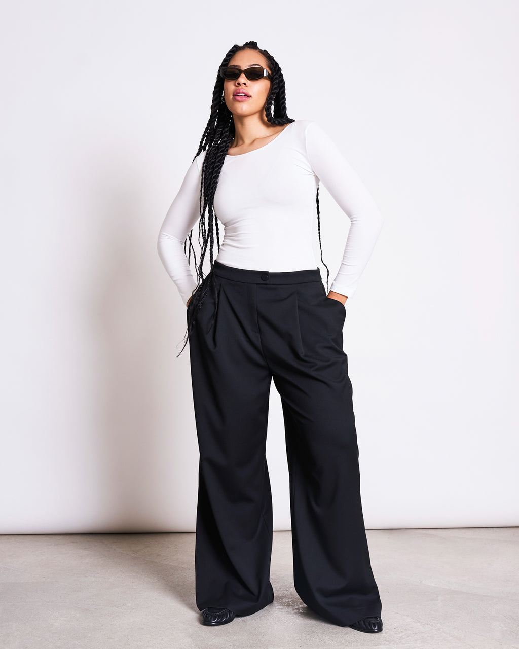 A woman with long braided hair wears sunglasses, loose black wide-leg trousers, black shoes, and the jan-n-june BALLERINA LONGSLEEVE LEON SOFT WHITE GOTS top, standing confidently with hands in pockets against a plain white background.