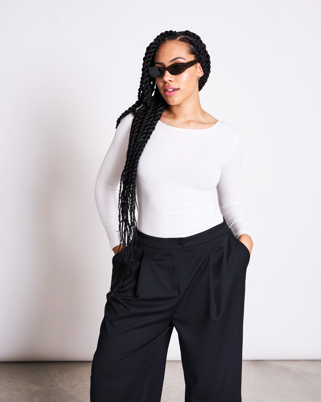 A woman with long braids and black sunglasses wears the jan-n-june BALLERINA LONGSLEEVE LEON SOFT WHITE GOTS top and loose black pants, standing against a plain light background with her hands in her pockets.