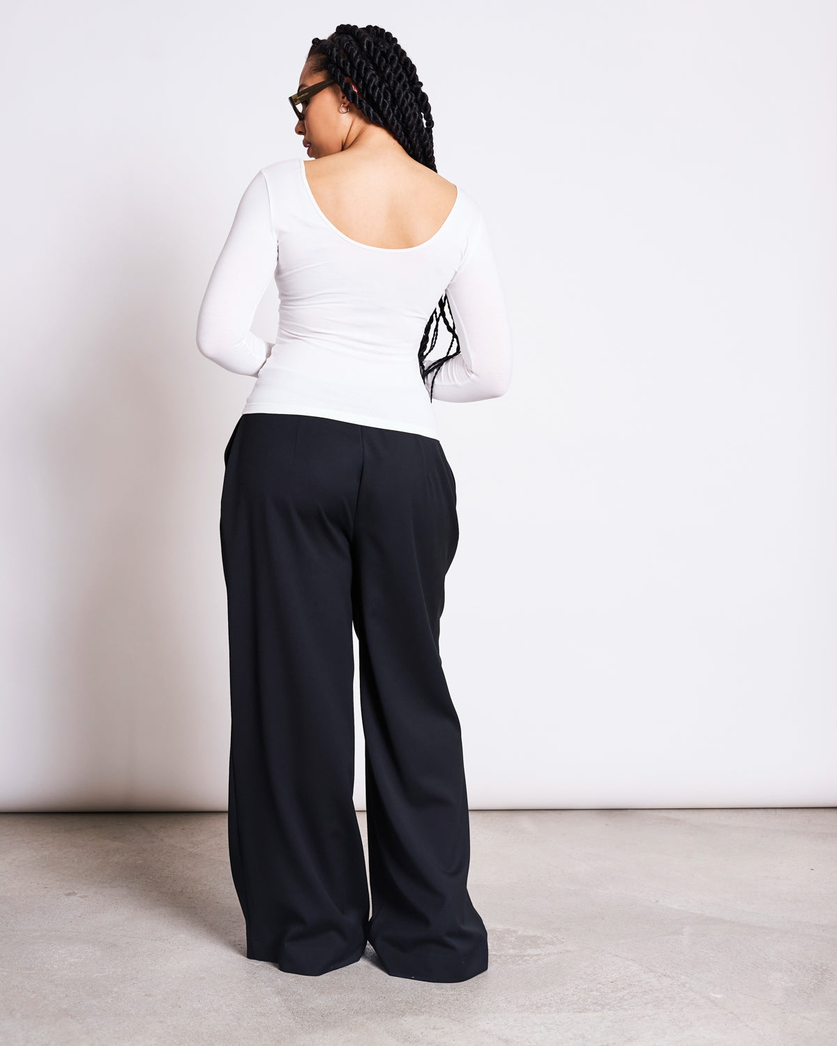 A person with long braids faces away from the camera, wearing jan-n-junes BALLERINA LONGSLEEVE LEON SOFT WHITE GOTS top and loose black pants, in a minimal indoor space with light walls and flooring.