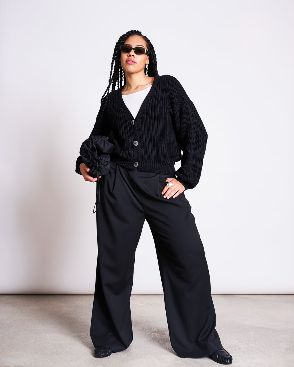 A woman stands confidently in front of a white wall wearing jan-n-junes CHUNKY CARDIGAN LENA BLACK GOTS over a white top, paired with wide-leg black pants, sunglasses, and holding a black bag.