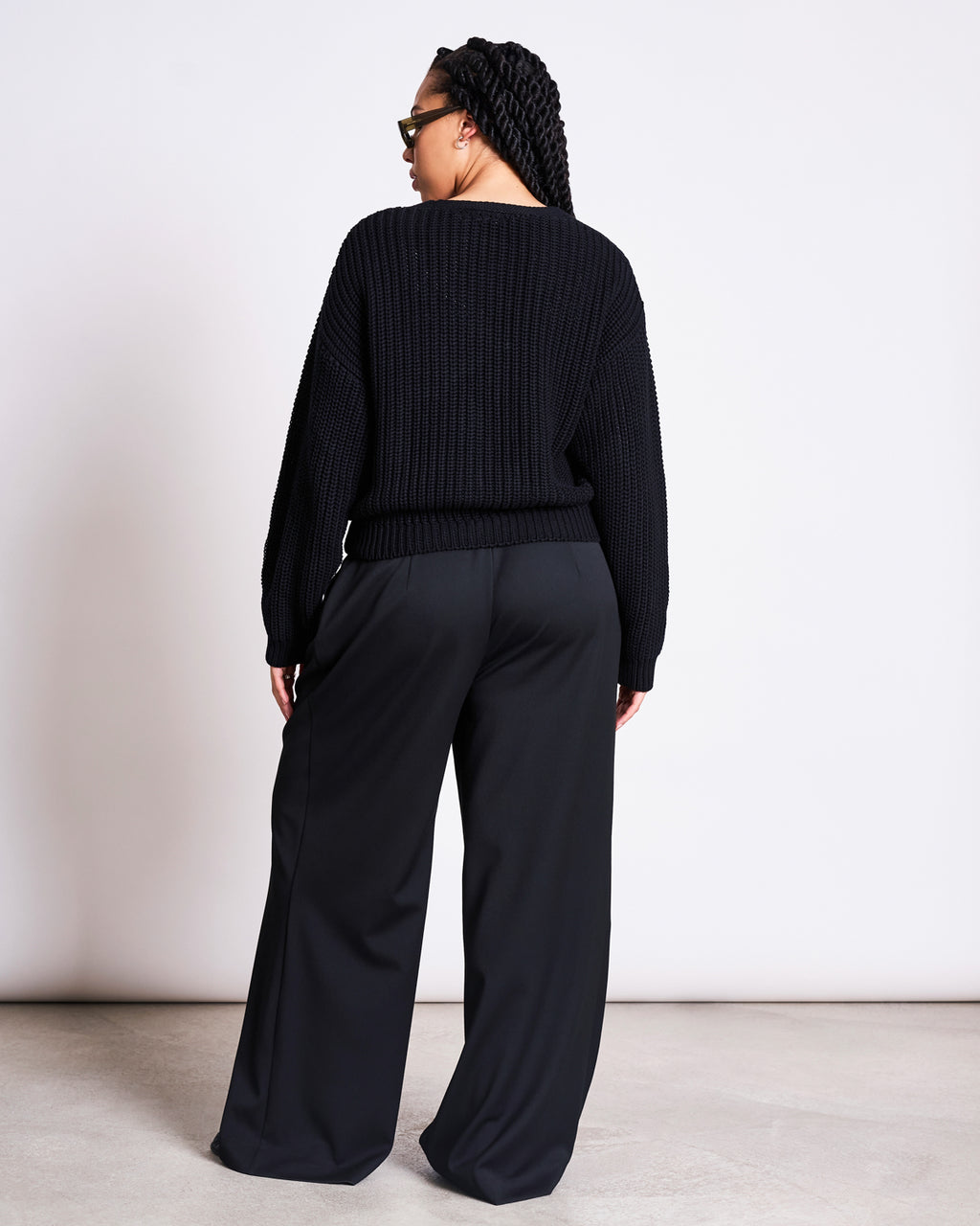 A person with long braided hair, seen from behind, wears the jan-n-june CHUNKY CARDIGAN LENA BLACK GOTS and wide-leg black pants against a plain white background.