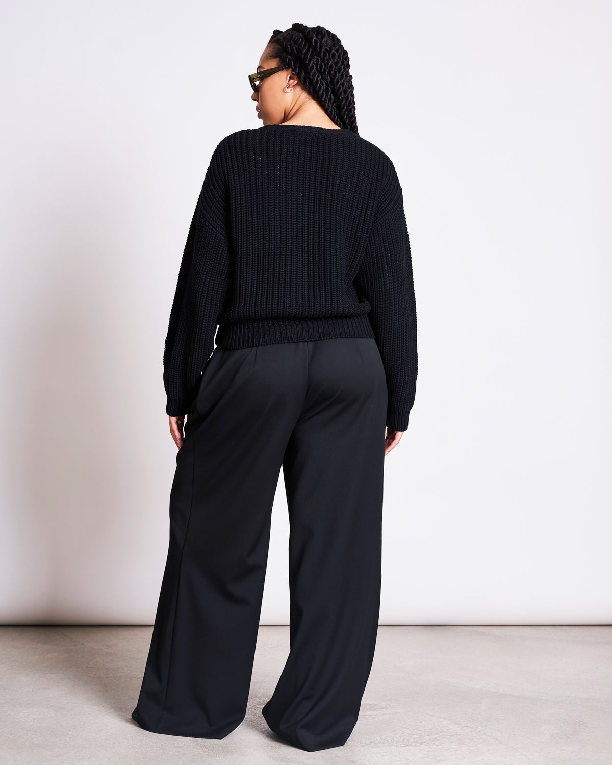 A person with long braided hair, seen from behind, wears the jan-n-june CHUNKY CARDIGAN LENA BLACK GOTS and wide-leg black pants against a plain white background.