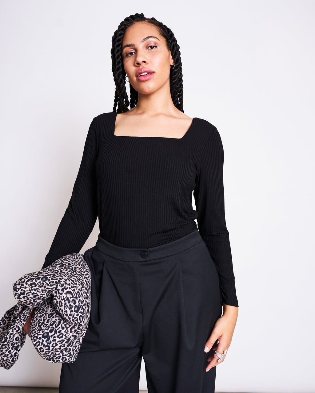 A woman with braided hair wears the jan-n-june SQUARE NECK LONGSLEEVE FRIA BLACK shirt with black trousers, holding a leopard print jacket in her left hand against a plain light background.