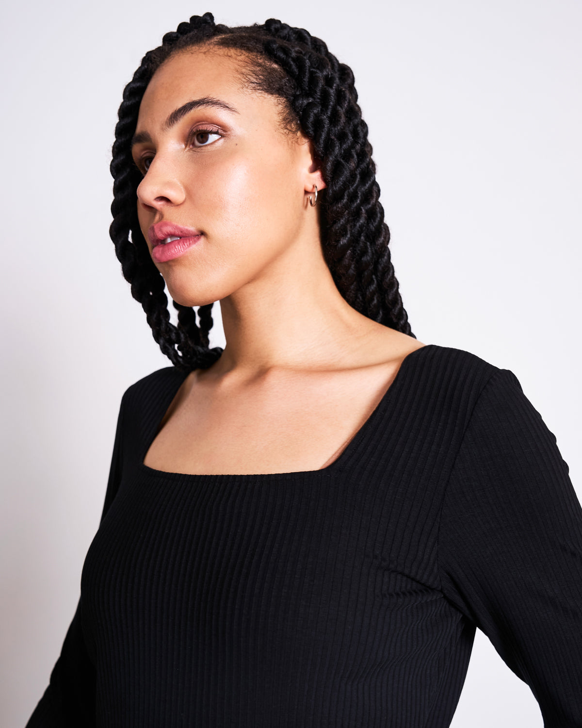 A woman with long braids wearing the jan-n-june SQUARE NECK LONGSLEEVE FRIA BLACK top and small hoop earrings gazes to the side, posed against a plain light background.