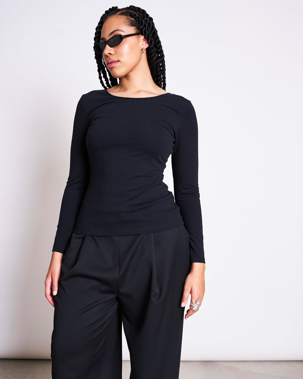 A woman with braided hair wears black sunglasses and the jan-n-june BALLERINA LONGSLEEVE LEON SOFT BLACK GOTS top with a boat neck, paired with loose black trousers. She stands against a plain white background, looking to the side.