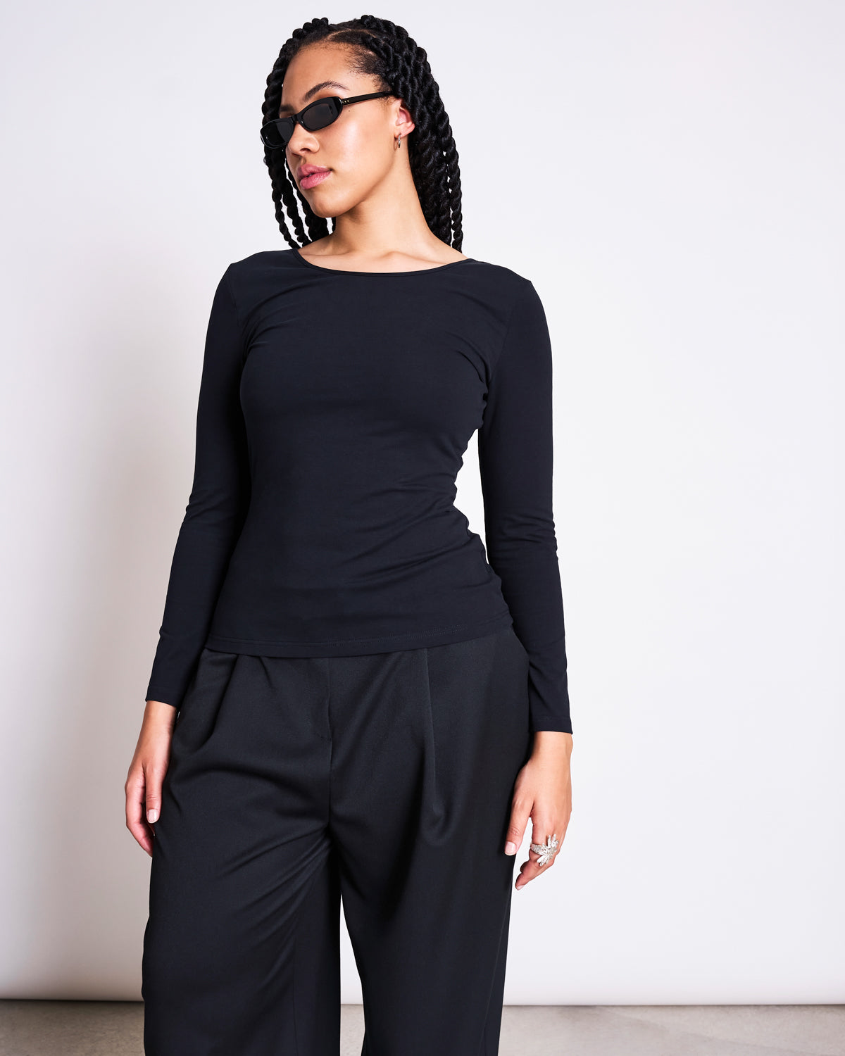 A woman with braided hair wears black sunglasses and the jan-n-june BALLERINA LONGSLEEVE LEON SOFT BLACK GOTS top with a boat neck, paired with loose black trousers. She stands against a plain white background, looking to the side.