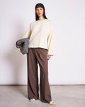 A woman stands against a plain background wearing the jan-n-june BOXY LONGSLEEVE ANKIRA BUTTERMILK top in an oversized fit, paired with wide-leg brown pants, sandals, a brown net headscarf, and holding a folded leopard-print garment.