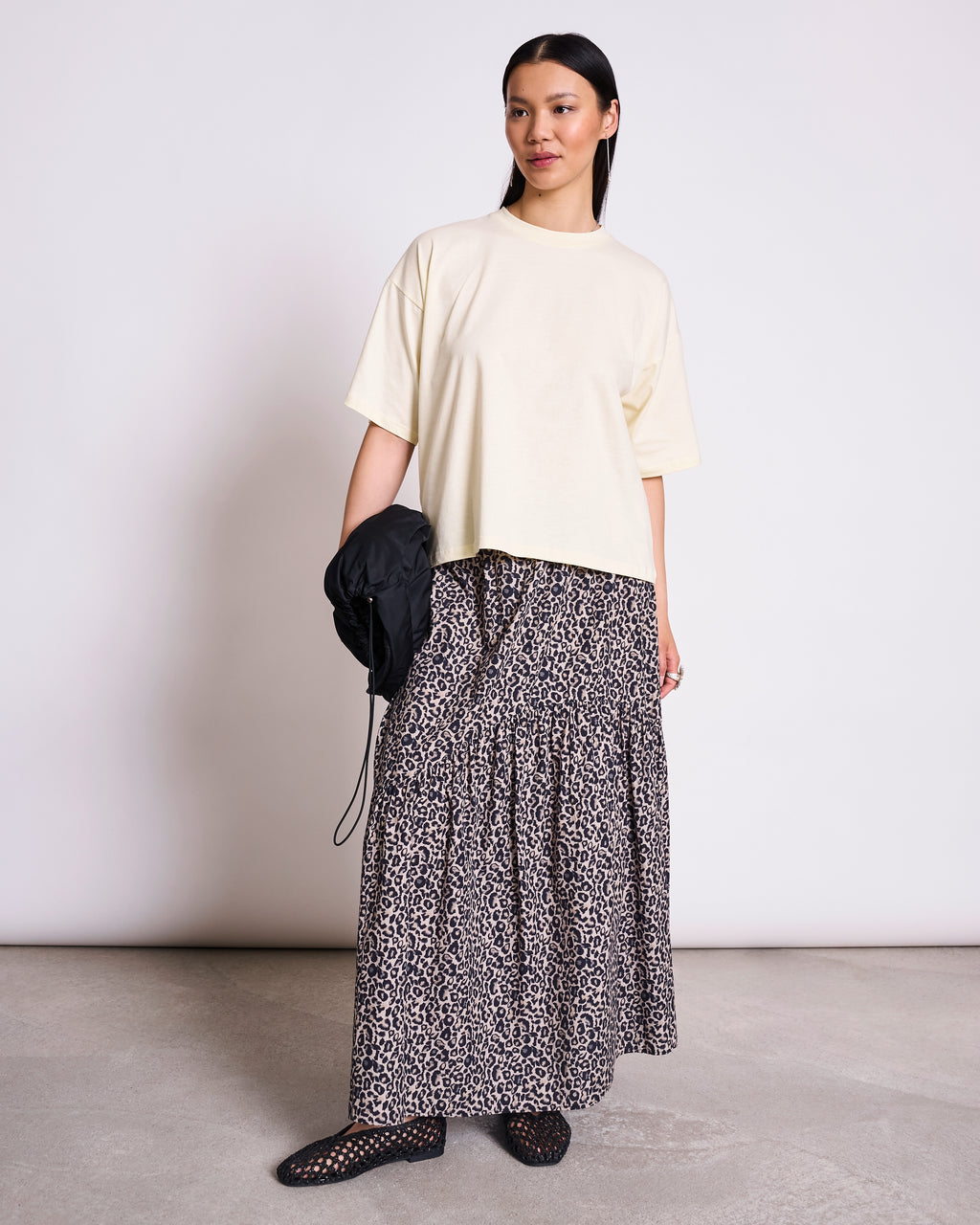 A woman with long dark hair and a calm expression wears the jan-n-june BOXY T-SHIRT TEMEL BUTTERMILK against a plain background, styled with a leopard print skirt, black woven shoes, and holding a black bag.