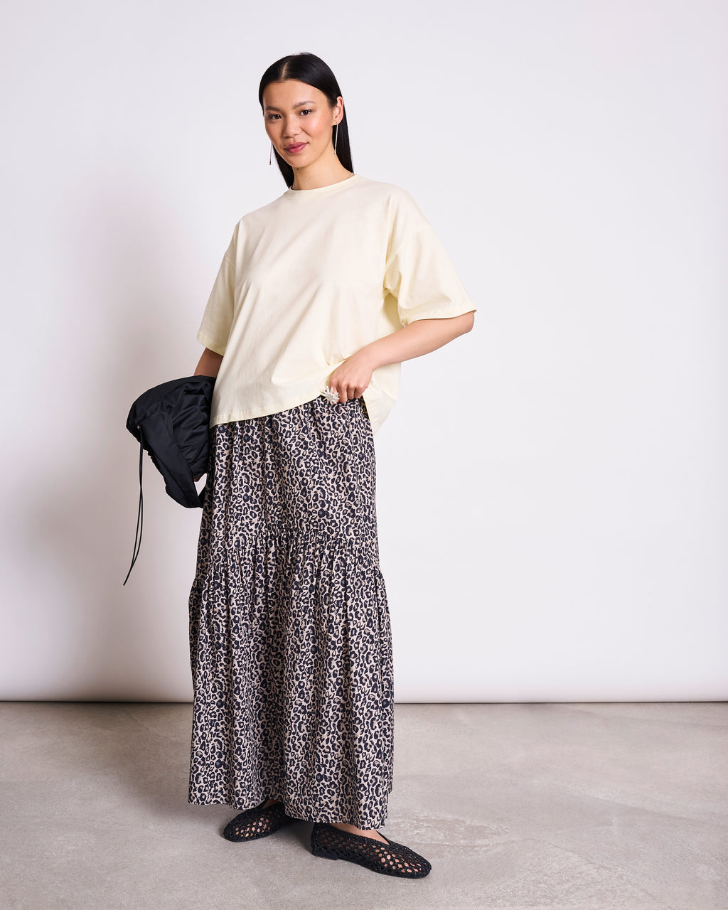 A woman stands against a plain background wearing the jan-n-june BOXY T-SHIRT TEMEL BUTTERMILK, an oversized cream top, paired with a long tiered leopard print skirt, black woven shoes, and holding a small black bag.