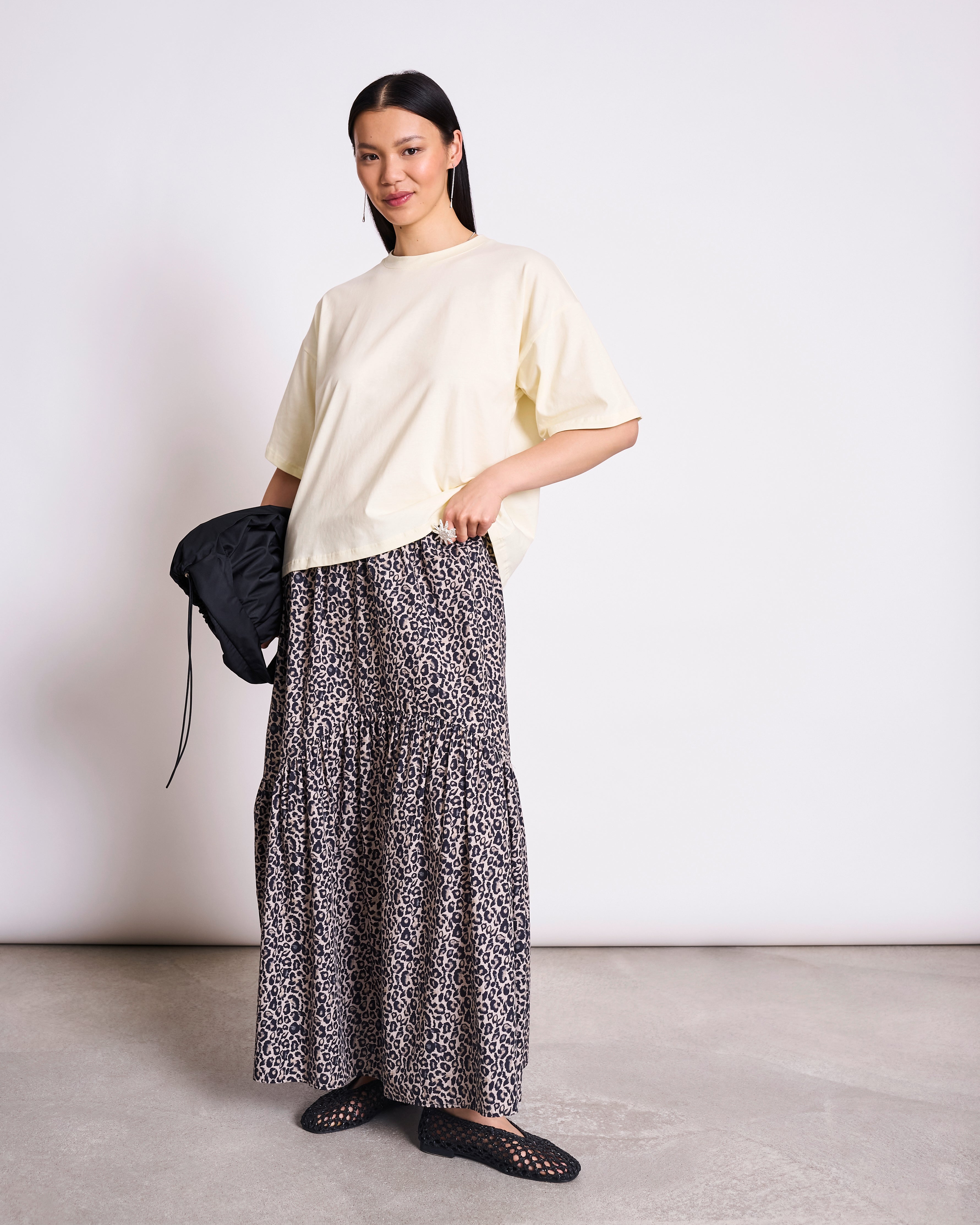 A woman stands against a plain background wearing the jan-n-june BOXY T-SHIRT TEMEL BUTTERMILK, an oversized cream top, paired with a long tiered leopard print skirt, black woven shoes, and holding a small black bag.