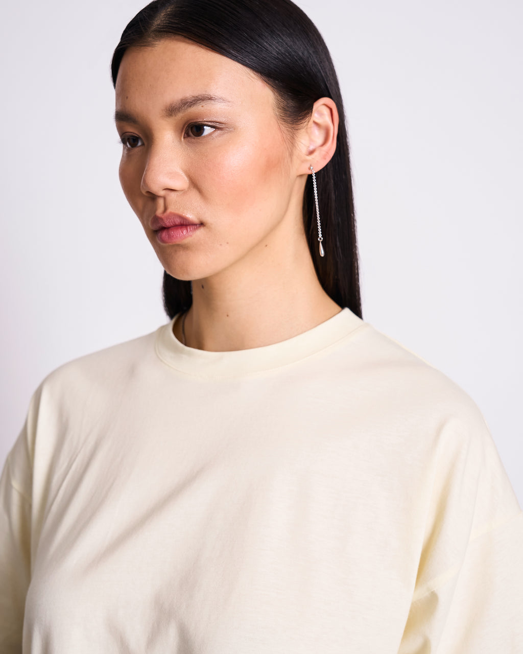 A woman with straight, dark hair wears the jan-n-june BOXY T-SHIRT TEMEL BUTTERMILK and a long, dangling earring. She looks slightly to the side with a neutral expression against a plain light background.