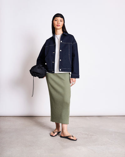 A woman stands against a white background wearing a dark denim jacket, white top, jan-n-junes MIDI KNIT SKIRT EBBA SEA SPRAY GOTS in olive green, black flip-flops, a black bandana, and holding a black bag at her side.