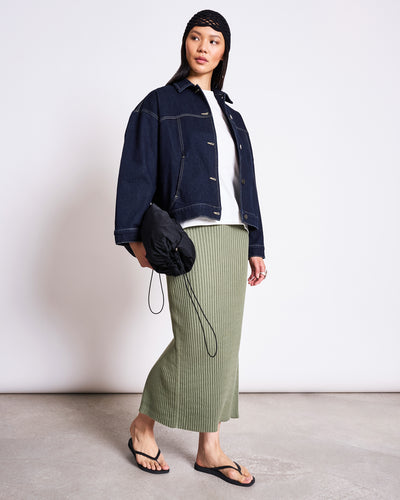 A woman in a black headscarf, dark denim jacket, white top, jan-n-june MIDI KNIT SKIRT EBBA SEA SPRAY GOTS, black flip-flops, and holding a black clutch stands against a plain background.