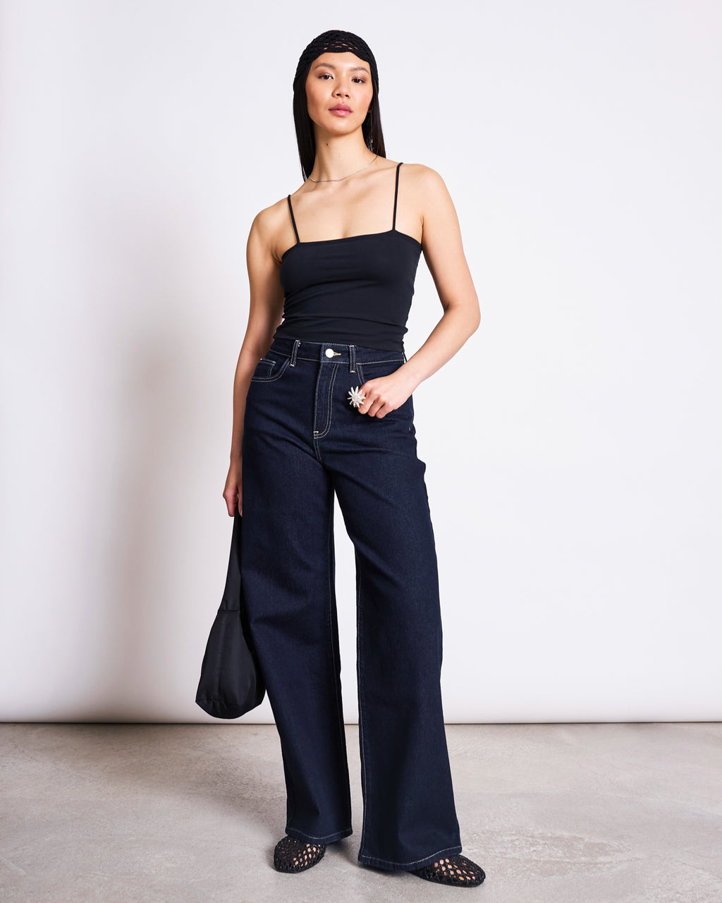 A woman poses with one hand in her pocket, wearing the jan-n-june TOP SOL BLACK GOTS organic cotton cropped top, styled with dark wide-leg jeans, black woven shoes, a black headscarf, and a black bag against a plain white wall.