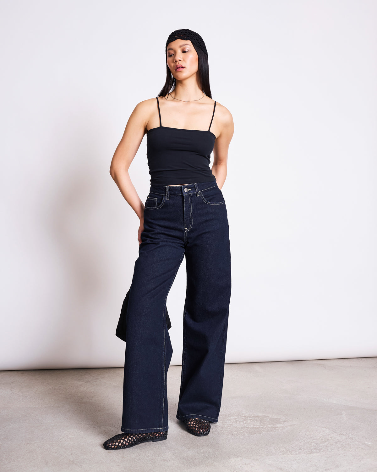 A woman stands against a plain white wall, wearing the jan-n-june TOP SOL BLACK GOTS spaghetti strap top, high-waisted wide-leg jeans, black studded shoes, and a black headband. Her arms are relaxed behind her back.