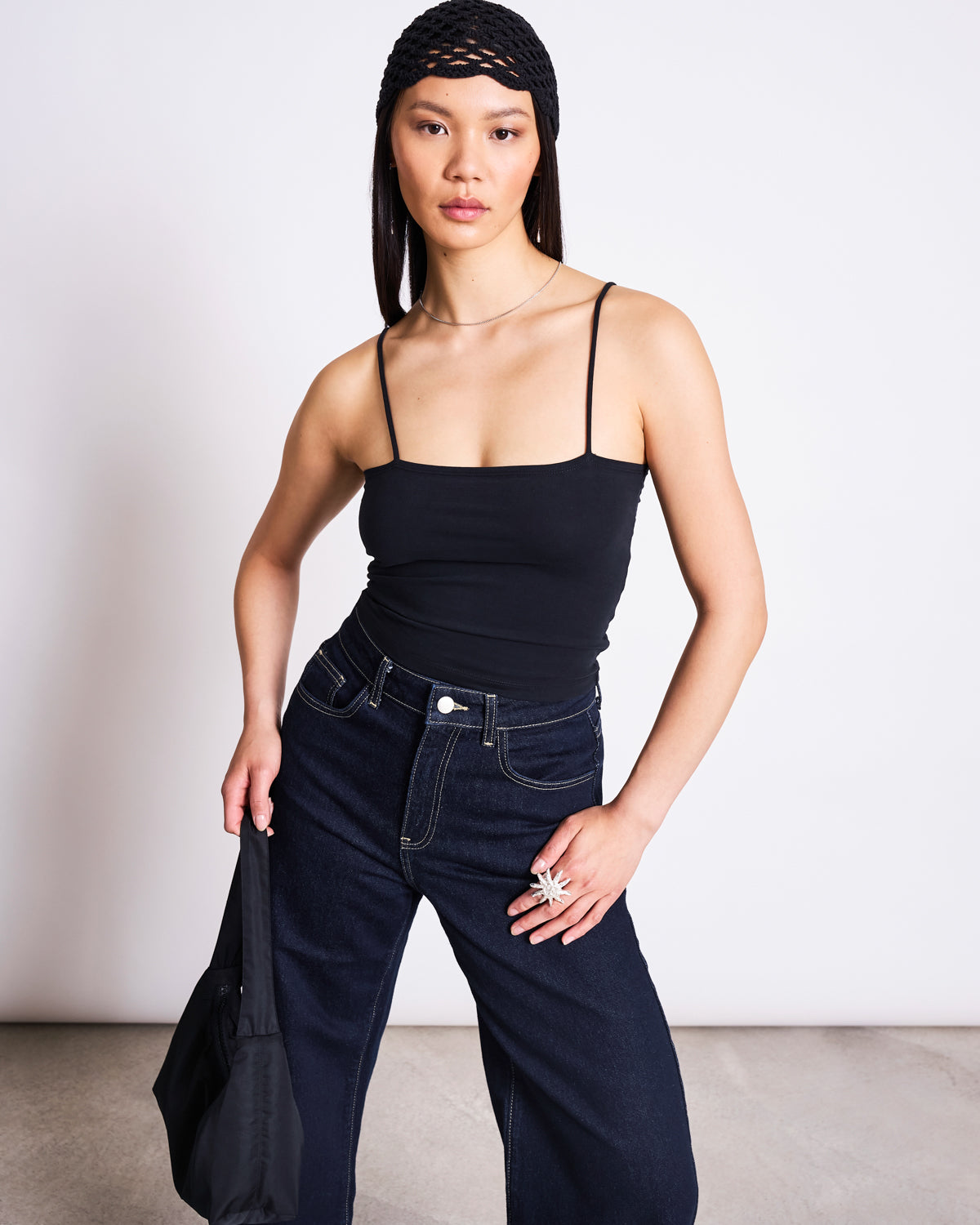 A woman poses with one hand on her hip, wearing the jan-n-june TOP SOL BLACK GOTS cropped organic cotton top, a black headband, dark wide-leg jeans, and holding a black bag against a plain background.