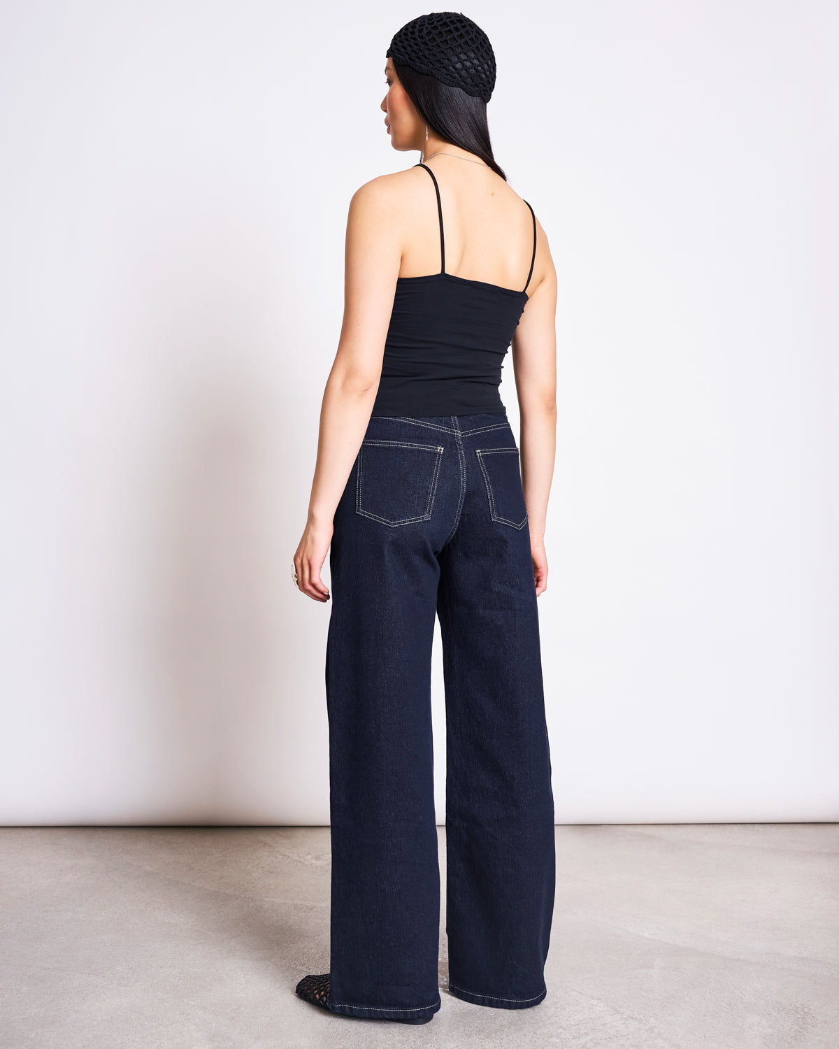 A woman faces away from the camera, wearing the jan-n-june TOP SOL BLACK GOTS organic cotton cropped top, wide-leg dark jeans with contrast stitching, a black crochet hat, and black shoes, against a plain white background.