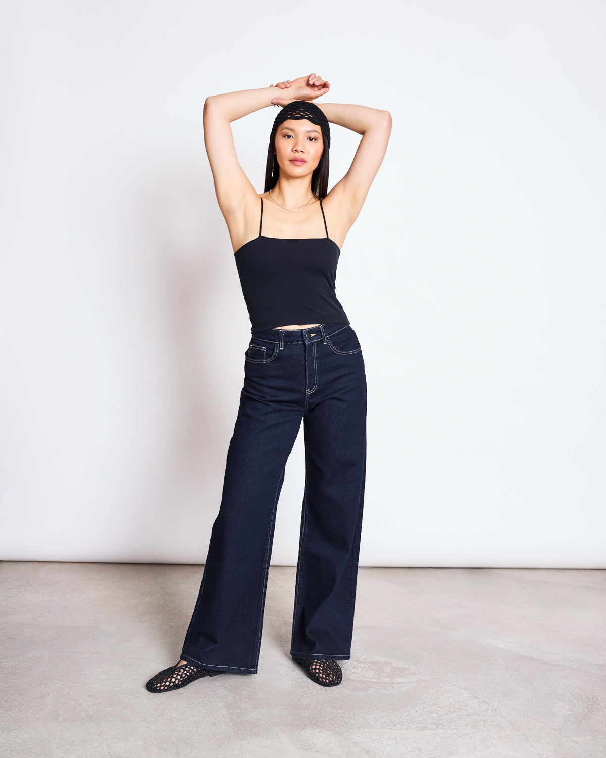 A woman stands with arms raised above her head, wearing the jan-n-june TOP SOL BLACK GOTS organic cotton cropped top, dark blue wide-leg jeans, black mesh shoes, and a black headband against a plain white background.