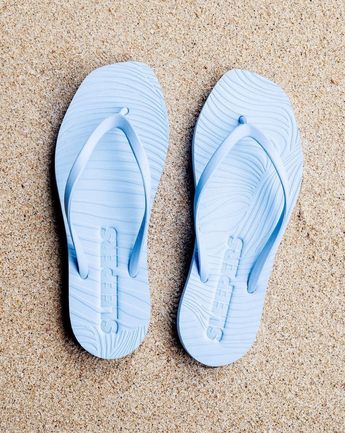 A pair of jan-n-june SLEEPERS TAPERED FLIP FLOPS in Angel Blue with a wavy textured pattern rest on sand, crafted from biodegradable materials for sustainable style.