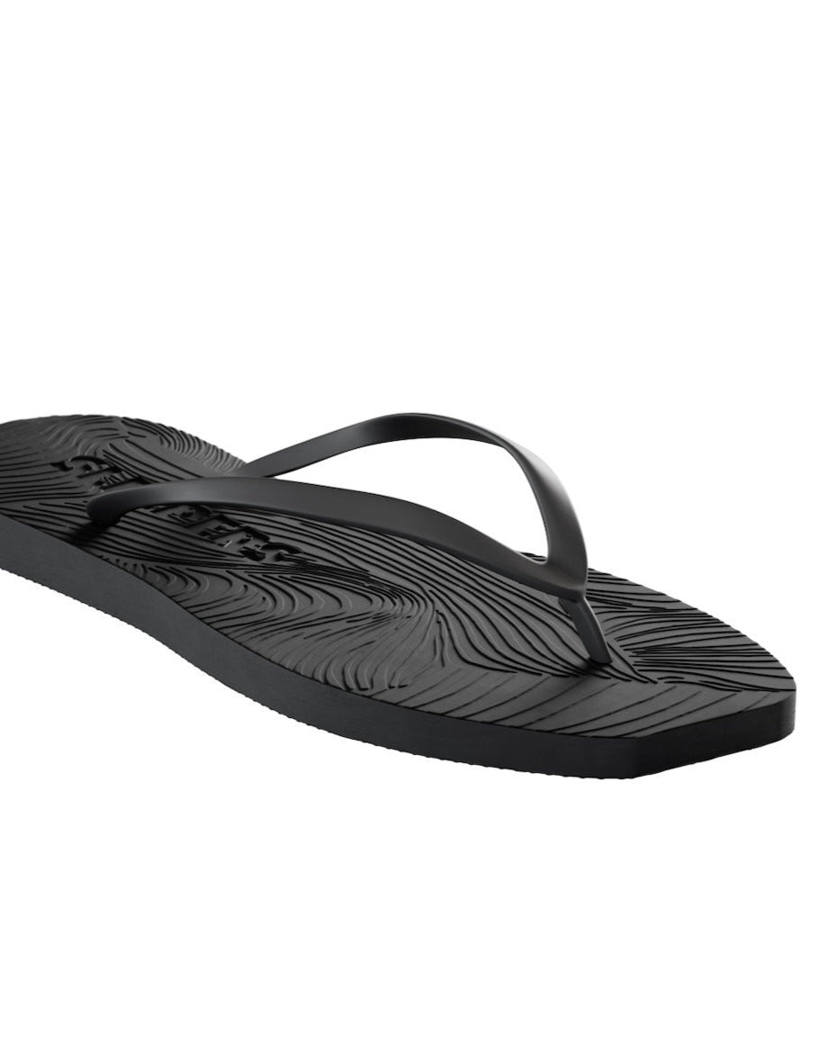 The jan-n-june SLEEPERS TAPERED FLIP FLOPS BLACK features a single black flip flop with textured, wavy patterns on the sole, a simple black thong strap, and sustainable design, shown against a white background.