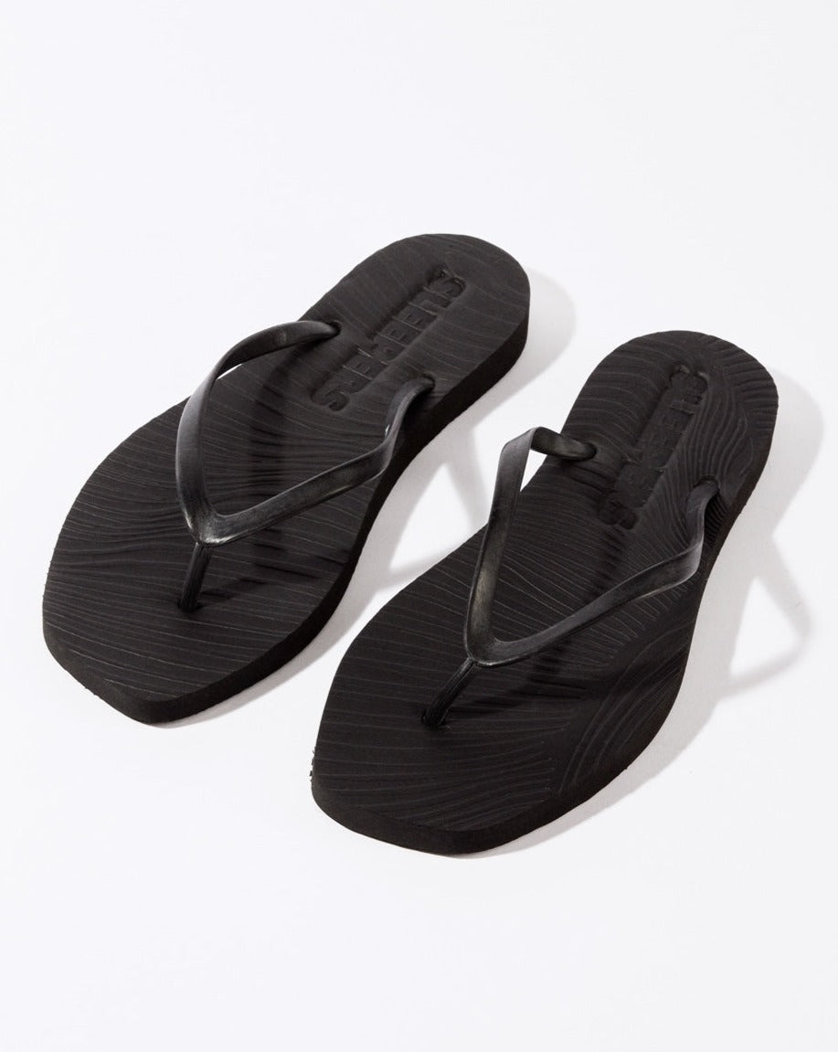 A pair of jan-n-june SLEEPERS TAPERED FLIP FLOPS BLACK, featuring thin straps and textured soles, made from biodegradable materials, are placed side by side on a white background.