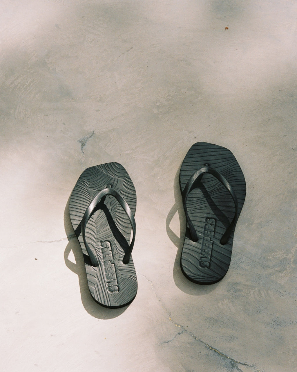 The jan-n-june SLEEPERS TAPERED FLIP FLOPS BLACK, featuring a woodgrain pattern, rest on light concrete in the sun—blending sustainable fashion and casual comfort.