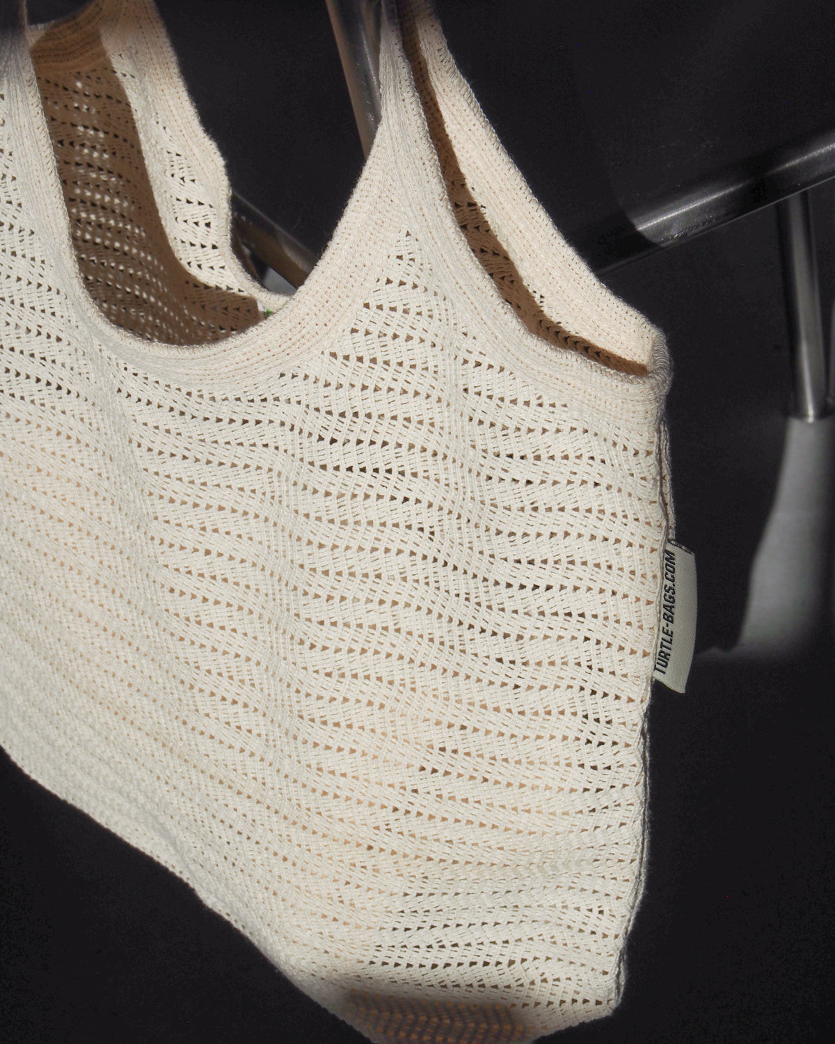 The ANTIQUE LACE BAG COTTON by jan-n-june is a cream, crocheted organic cotton bag with a woven texture and side label, shown hanging on a clear chair in soft light. It also serves as a reusable shopping bag.
