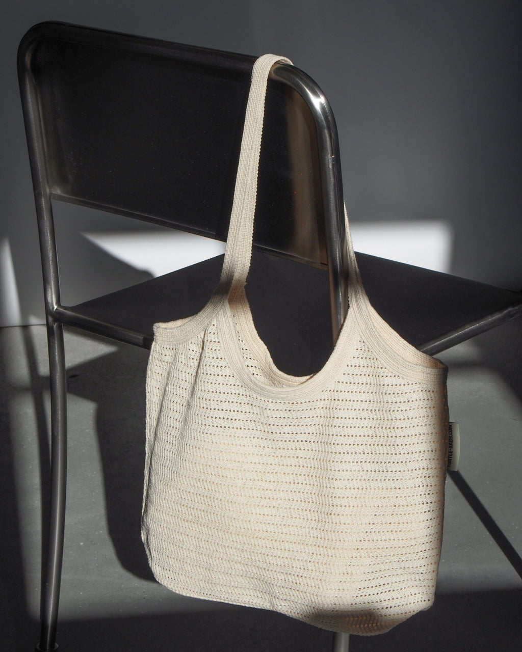 The ANTIQUE LACE BAG COTTON by jan-n-june, a beige knitted organic cotton bag with a wide strap, hangs on the back of a metal chair as sunlight casts shadows across the gray floor and chair.