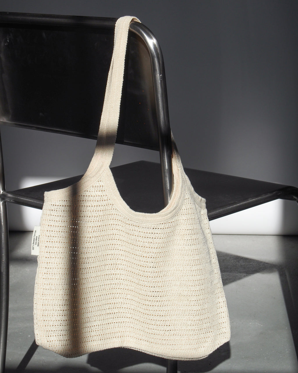 The ANTIQUE LACE BAG COTTON by jan-n-june, a fair trade cream crochet bag made from organic cotton, hangs on a black metal chair’s backrest and casts gentle shadows in bright natural light.