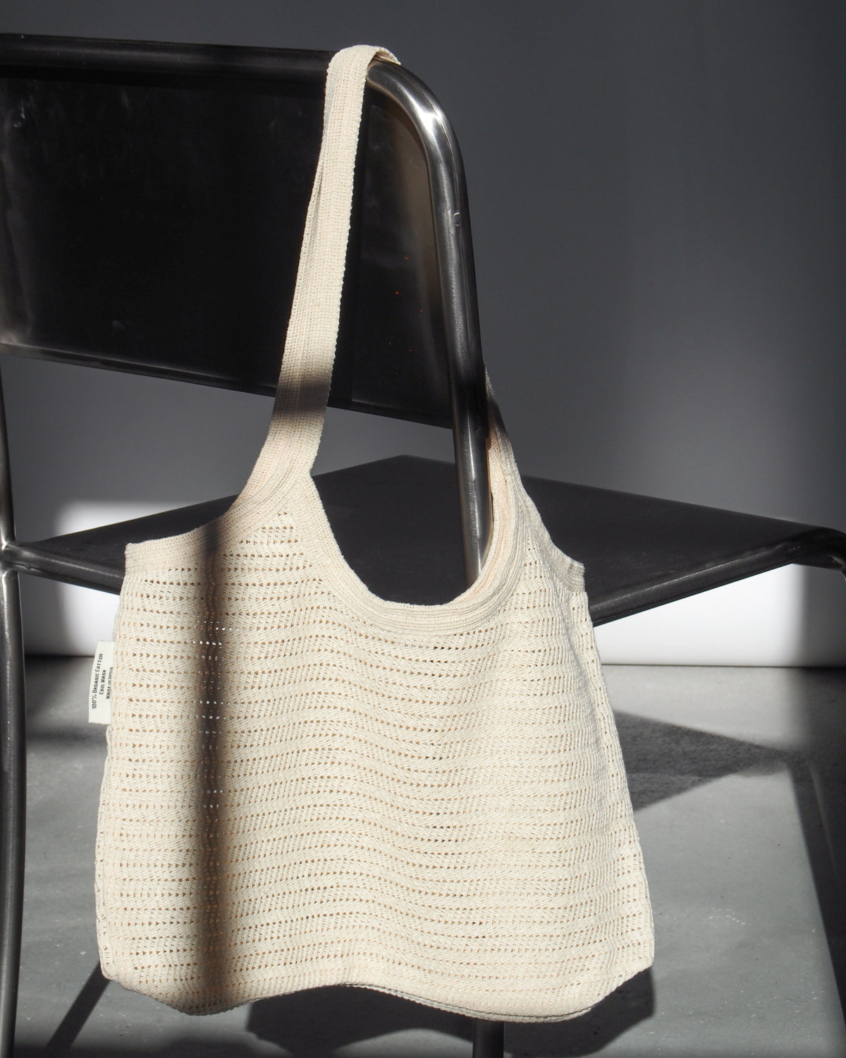 The ANTIQUE LACE BAG COTTON by jan-n-june, a fair trade cream crochet bag made from organic cotton, hangs on a black metal chair’s backrest and casts gentle shadows in bright natural light.