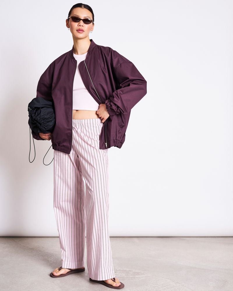 A person stands confidently against a white background wearing jan-n-june’s OVERSIZED BLOUSON TANARO CHERRY LACQUER, sunglasses, a white crop top, pink and white striped pants, sandals, and holding a black bag.