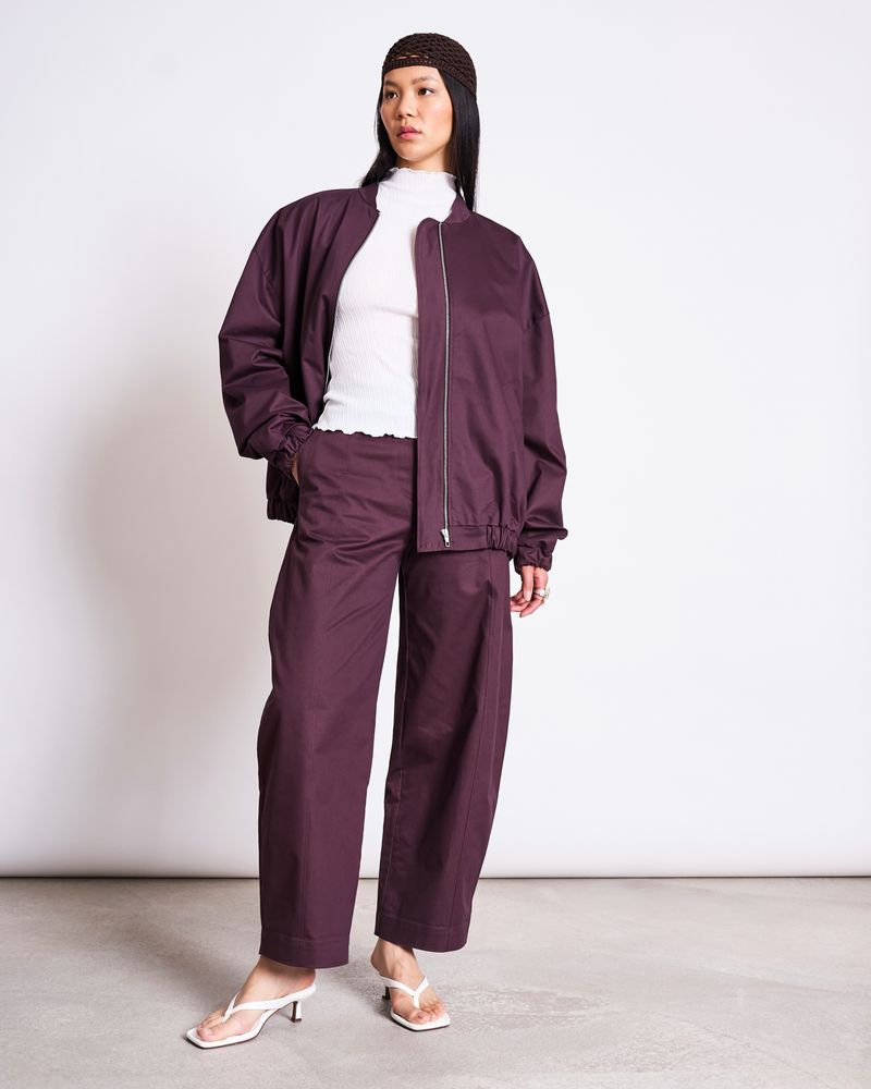 A woman stands against a plain background wearing the jan-n-june OVERSIZED BLOUSON TANARO CHERRY LACQUER with matching wide-leg pants, a white ribbed turtleneck, white heeled sandals, and a dark brown headband.