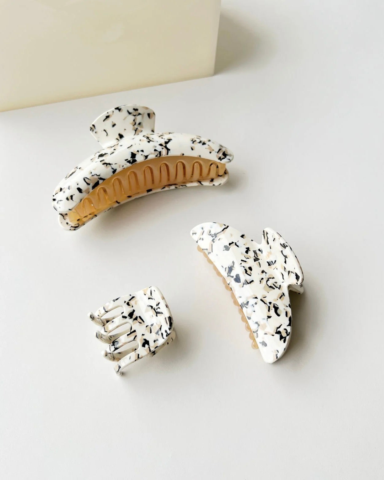 Three speckled white and black MIDI HEIRLOOM CLAW TERRAZZO clips by jan-n-june, in varying sizes, are arranged on a light surface, with a beige rectangular object partially visible in the background.
